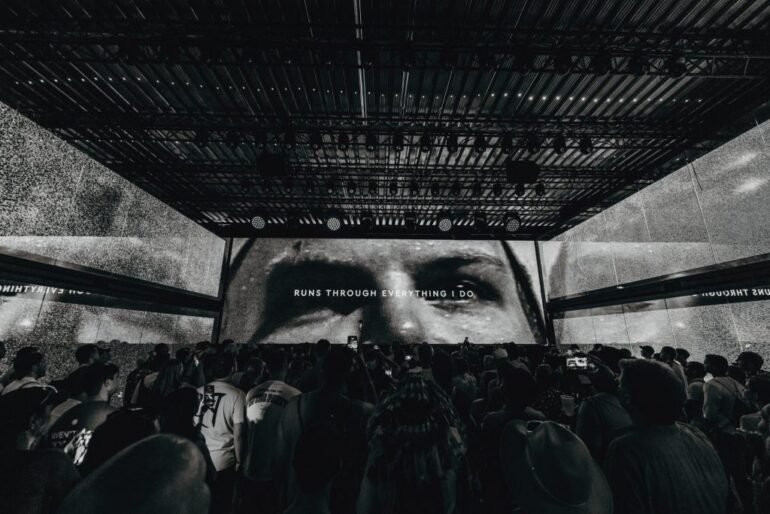 Avicii Tomorrowland 2026 tendrá un homenaje único | Pulsar Mix Radio FM Large crowd at a concert in a dark venue, with a massive screen showing'RUNS THROUGH EVERYTHING I DO'.