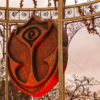 Rusty shield sculpture with a stylized rose design hanging inside ornate metal arches outdoors.