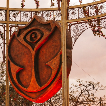 Rusty shield sculpture with a stylized rose design hanging inside ornate metal arches outdoors.