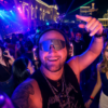 Man in front with oversized headphones and a visor, smiling at a crowded neon-lit night party as others dance around him