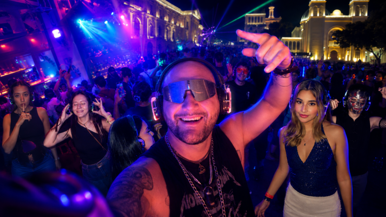 Man in front with oversized headphones and a visor, smiling at a crowded neon-lit night party as others dance around him